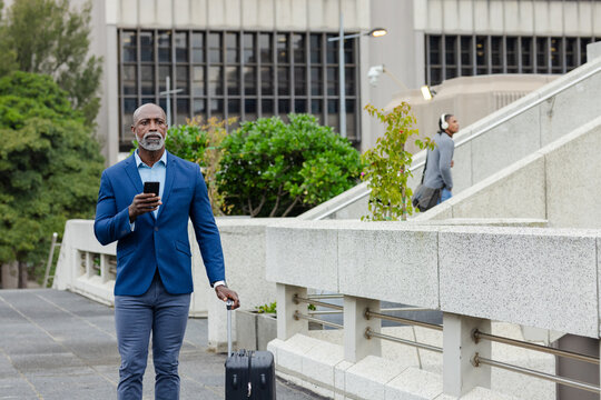 African american man wearing blue blazer walking along plaza pulling suitcase, checking smartphone