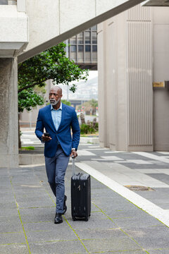 African american man walking across plaza wearing navy blazer, pulling suitcase, holding smartphone