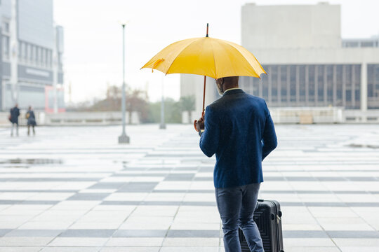 African american man walking across wet plaza pulling black suitcase under umbrella toward office