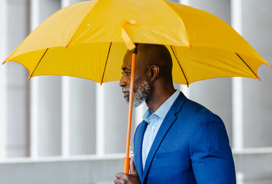 Mature african american man in blazer holding umbrella walking outside office building