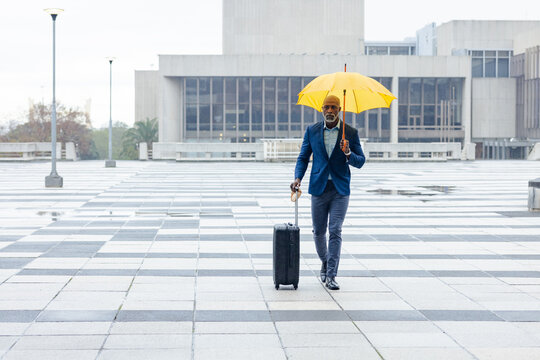 Senior african american man walking across plaza with puddles holding umbrella and black suitcase