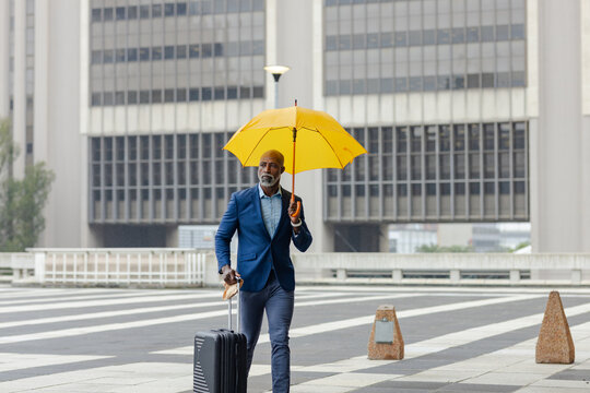 Senior african american man crossing striped crosswalk holding yellow umbrella with black suitcase