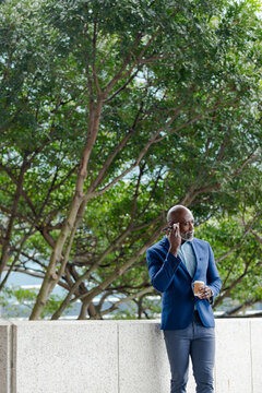 African american businessman leaning on plaza wall talking on phone holding coffee cup