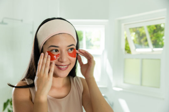 Asian woman applying red under-eye gel patches in bathroom, wearing pink headband and beige top