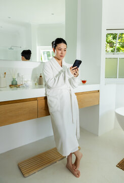 Asian woman in robe checking smartphone after alert, reading message in bathroom at wooden vanity
