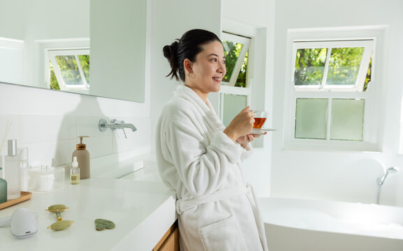 Adult Asian woman standing in white plush bathrobe in home bathroom, holding clear glass teacup