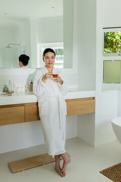 Woman standing in modern bathroom wearing white bathrobe, holding glass teacup and saucer