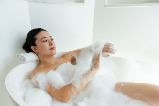 African American woman reclining in oval tub resting head on rolled towel lathering arm with foam