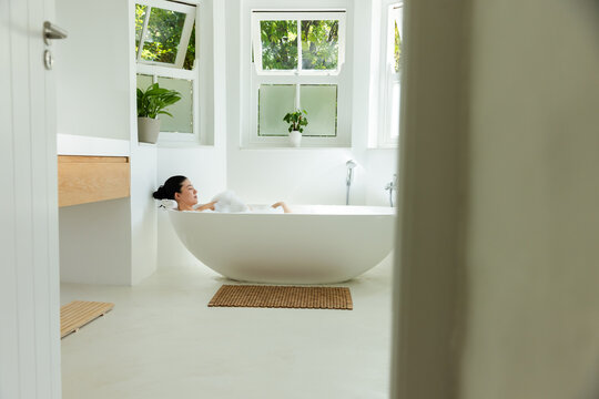 African American woman reclining in freestanding oval tub in bright bathroom wearing white towel