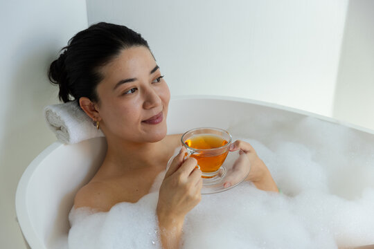 Woman reclining in white bathtub with thick foam, resting head on rolled towel, holding teacup