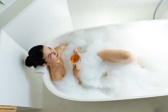 African American woman reclining in white tub with thick foam, holding glass cup with amber drink