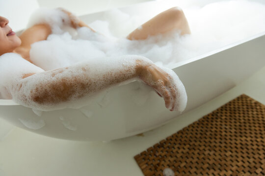 Adult female reclining in freestanding white bathtub, soaking in dense bath foam near wooden mat