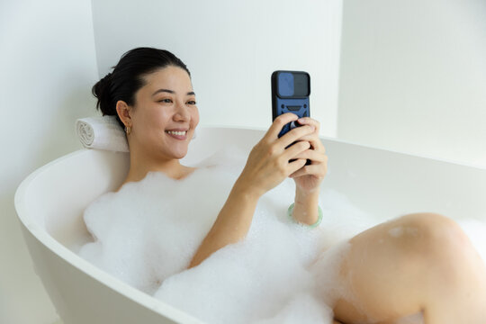 African American woman reclining in freestanding tub, being covered by foam, holding smartphone