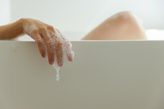 Adult soaking in bathroom, resting soapy hand over white bathtub rim with blurred knee and drip