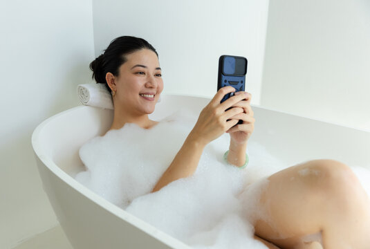 Asian woman reclining in tub in bathroom, holding phone in foam, wearing hoops and green bangle