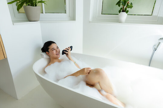 African American woman reclining in bathtub on rolled towel, holding smartphone with foam