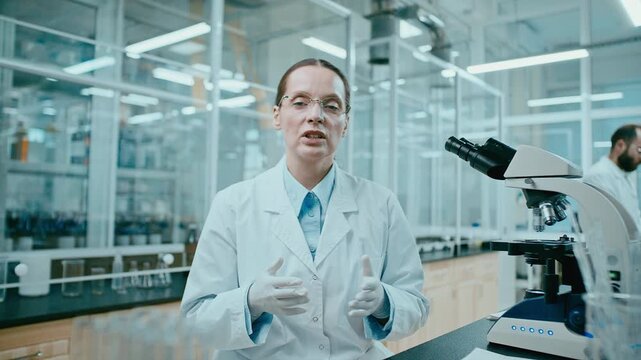Female scientist in lab coat and gloves looking at camera and explaining research findings while sitting near microscope in modern laboratory, having video call or filming vlog
