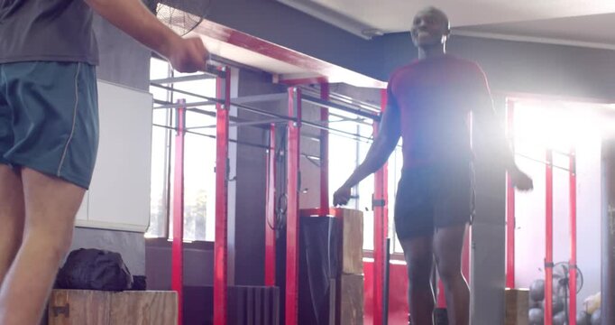Diverse male duo holding jump rope overhead and synchronizing jumps in gym for cardio workout