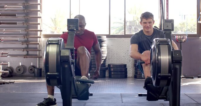 Diverse male training partners entering rowing machines, securing straps, syncing strokes at gym