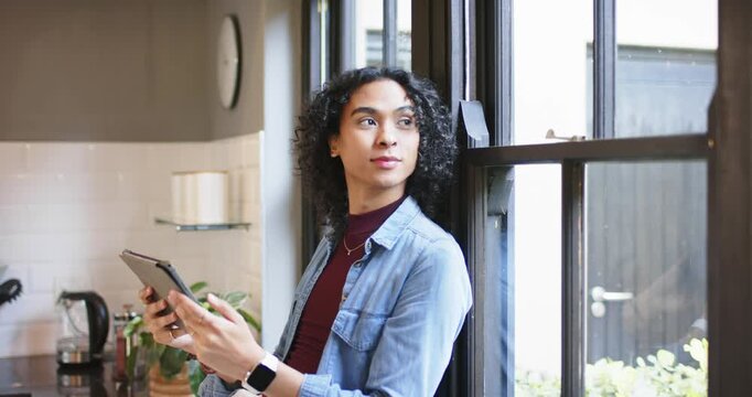 Non-binary individual tapping tablet with smartwatch at kitchen window gazing out then tucking hair