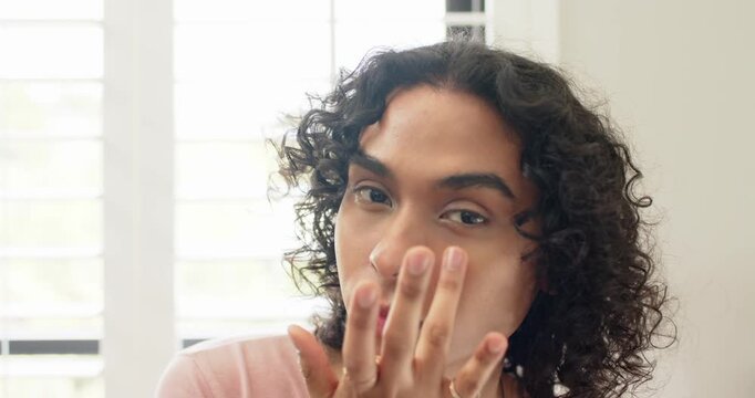 Non-binary adult looking at camera and blending cream by white shutters for skincare routine
