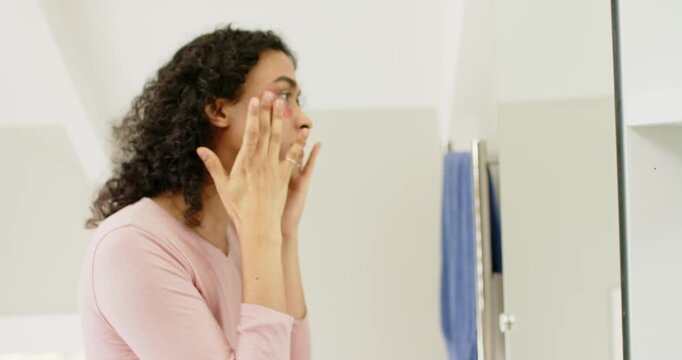 Holding patch and smoothing under right eye, non-binary person treating skin at bathroom mirror