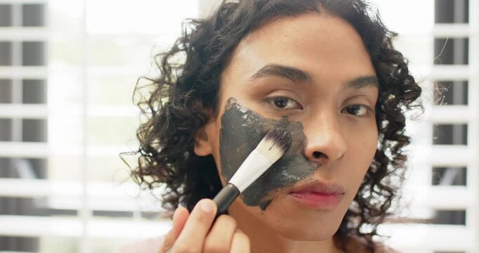Lifting brush toward face, non-binary adult applying charcoal mask across cheek at vanity
