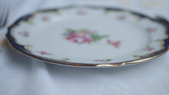 Close up cinematic footage of a white porcelain plate with a floral pattern and golden rim on a white tablecloth, slow pan reveals a fork beside it