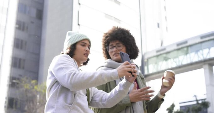 Female friends at plaza seeking directions on smartphone, pointing at skybridge, holding coffee