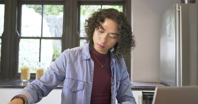 Non-binary person picking tote bag, writing notes, scrolling laptop and packing at kitchen table