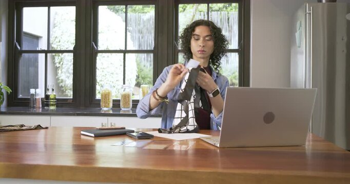 Non-binary adult picking up grid cloth in kitchen, unfolding design, noting, typing on laptop