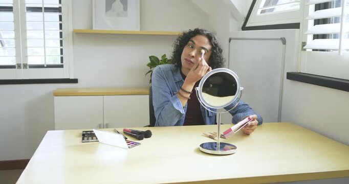 Non-binary adult using mirror, blending powder under eyes using palette and brushes at attic desk