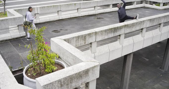 African American man stretching hamstring on roof as walker carrying smartphone passes, copy space