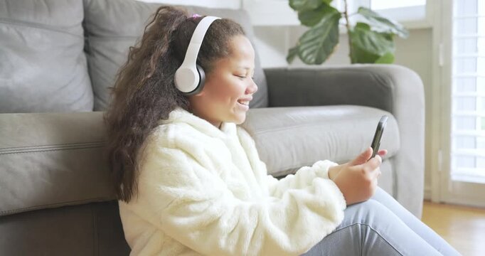 Preteen girl grasping smartphone with headphones leaning against sofa tapping screen in living room