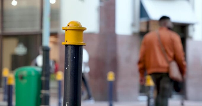 Diverse pedestrians starting walking to shops, man in brown jacket passing bollard with yellow cap