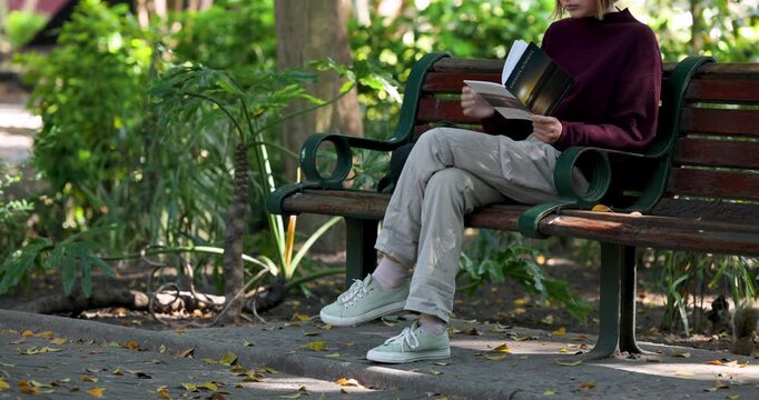 Reader is reading paperback, turning pages and adjusting book on green bench as pedestrians pass