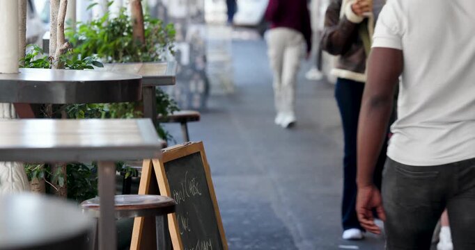 Passersby moving past cafe keeping chalk sign sharp while woman in shearling holding coffee passing