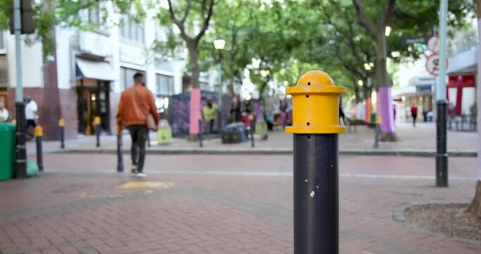 Diverse pedestrians walking across brick street, passing centered black bollard toward storefronts