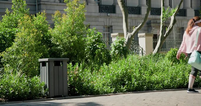 Pedestrians are entering from left on walkway, walking past gray trash bin, heading to class