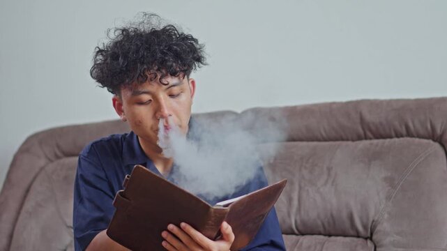 Focused young man using electronic cigarette while studying or journaling in leather book
