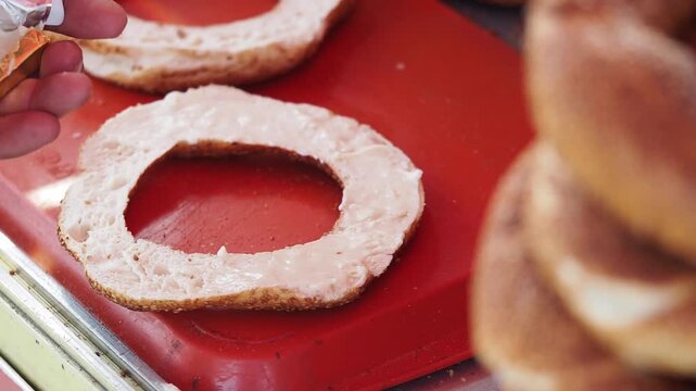 Street food vendor spreading soft cream cheese on fresh sesame simit bread on red tray