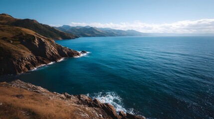 Fototapeta premium A breathtaking panoramic view of a rugged wild coastline meeting the deep blue ocean with distant mountains under a clear sunny sky
