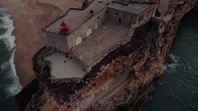 Drone orbit around nazar&eacute; lighthouse and forte de s&atilde;o miguel arcanjo, revealing cliff edge, stone architecture and Atlantic Ocean below in nazar&eacute; portugal big wave coastline