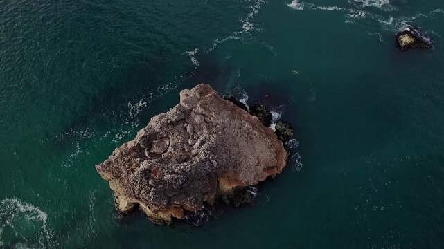 Top-down spinning drone shot over isolated sea stack near nazar&eacute; portugal, revealing textured rock surface, surrounding turquoise Atlantic water and circular motion emphasizing coastal geology