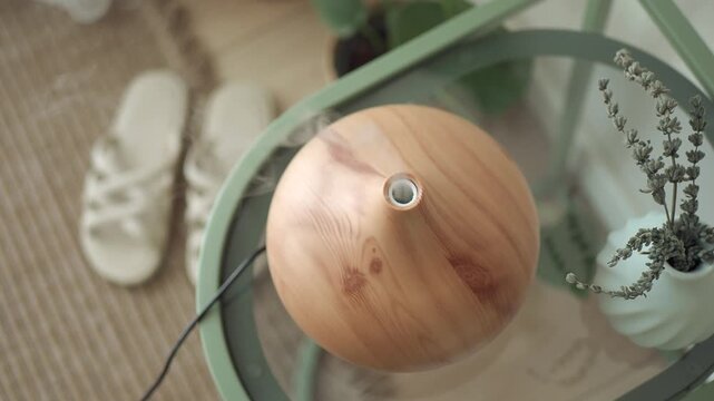 Aroma oil diffuser emitting steam on glass table near lavender and slippers in cozy room