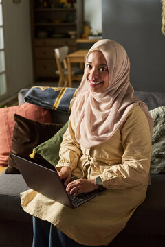 Portrait of young adult Muslim woman smiling while sitting on sofa using laptop, hands on keyboard, wearing hijab, looking into camera in home living room setting