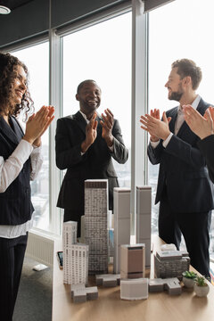 A diverse group of professionals in suits celebrates a successful project with applause near a window overlooking a cityscape, with architectural models on a table