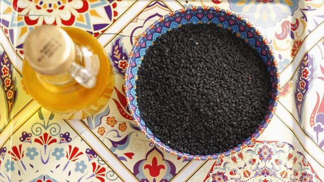 A close up top-down view of black cumin seeds and a bottle of nigella sativa oil, revealing traditional patterns on the background