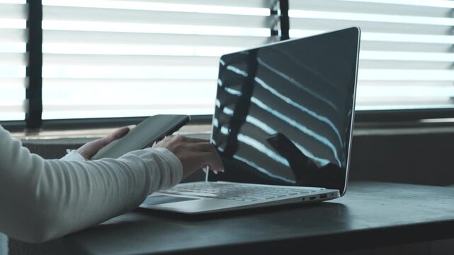 Close-up of a person using a smartphone beside a laptop on a desk, natural light highlighting a modern workspace and multitasking digital lifestyle.