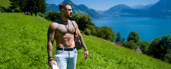 Naklejka premium Travel portrait with muscular man hiking mountain path. Travel adventure with confident man near alpine lake. Travel lifestyle with active hiker walking meadow. Travel inspiration in summer nature.